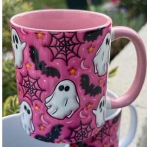 May include: A pink ceramic mug with a Halloween design. The mug has a repeating pattern of white ghosts, black bats, spiderwebs, stars, and moons. The inside and handle are also pink.