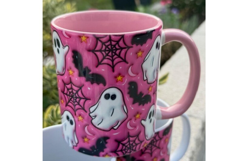 May include: A pink ceramic mug with a Halloween design. The mug has a repeating pattern of white ghosts, black bats, spiderwebs, stars, and moons. The inside and handle are also pink.