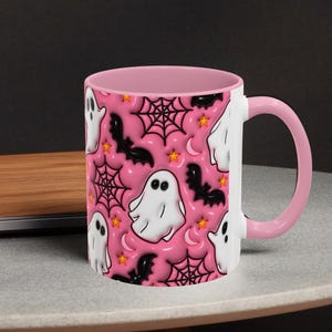 May include: A white ceramic mug with a pink interior and handle. The mug features a repeating pattern of white ghosts, black bats, spiderwebs, stars, and crescent moons on a pink background. The mug is sitting on a grey surface.