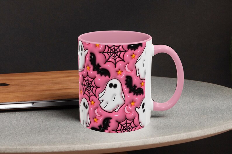 May include: A white ceramic mug with a pink interior and handle. The mug features a repeating pattern of white ghosts, black bats, spiderwebs, stars, and crescent moons on a pink background. The mug is sitting on a grey surface.