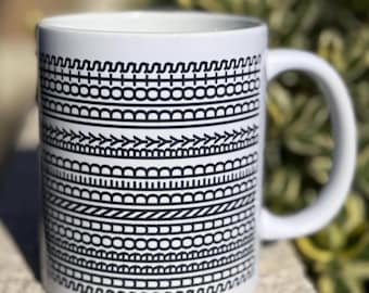 May include: White ceramic mug with a black geometric pattern. The design features various horizontal bands of repeating shapes, including wavy lines, circles, and arrows. The mug has a curved handle and is set against a blurred green background.