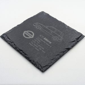 May include: A black slate coaster featuring a white outline of a Nissan K11 Micra car. The coaster is engraved with the text "K11 Micra", "1.3 Litre NA Inline 4", "74 bhp", "103 Nm", "0-62: 12 seconds", "Top Speed: 106 mph", and "Made in Japan".