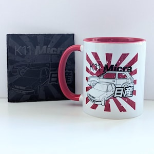 May include: White ceramic mug with a red handle and interior, displaying a K11 Micra car graphic with Japanese text against a red sunburst. A black slate coaster with a car graphic and text is also visible.