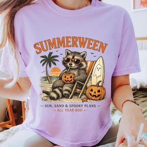 May include: A light purple t-shirt featuring a graphic of a raccoon wearing sunglasses, sitting in a beach chair, holding a jack-o'-lantern drink. The text reads SUMMERWEEN, SUN, SAND & SPOOKY PLANS, and ALL YEAR BOO, with a surfboard and palm tree.