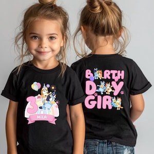 A young girl wearing a black t-shirt with a birthday theme, featuring the character Bluey from the children's television show Bluey's Friends.