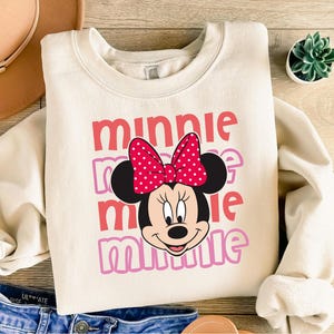 A white sweatshirt with a Minnie Mouse design printed on it, along with a pair of jeans and a hat.
