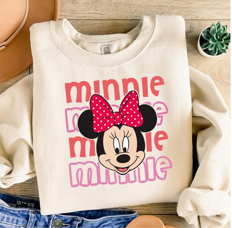 A white sweatshirt with a Minnie Mouse design printed on it, along with a pair of jeans and a hat.
