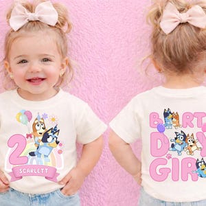 A smiling young girl wearing a white t-shirt with a birthday theme, featuring characters from the children's television show "Bluey and Friends".