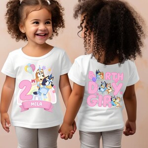 A young girl wearing a white t-shirt with a cartoon character design and the text "2 MILA" printed on it.