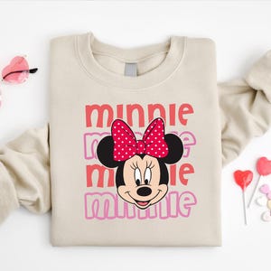 A beige sweatshirt with a Minnie Mouse design and the text "Minnie Minnie" printed on it, along with a pair of pink sunglasses and heart-shaped lollipops.