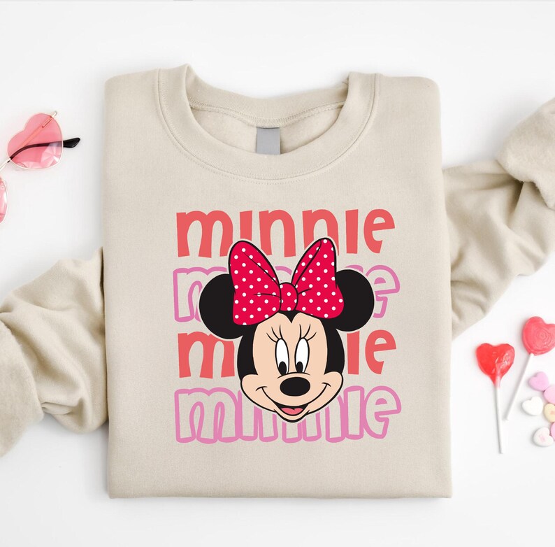 A beige sweatshirt with a Minnie Mouse design and the text "Minnie Minnie" printed on it, along with a pair of pink sunglasses and heart-shaped lollipops.