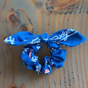 Puede incluir: Dos coleteros azules con texto blanco y detalles rojos. Uno está atado en forma de lazo, el otro es un coletero estándar. Están sobre una superficie de madera.