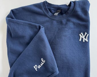 May include: Navy blue sweatshirt with the New York Yankees logo embroidered on the chest. The name "Paul" is embroidered on the left sleeve in white thread. The sweatshirt is folded on a white surface.