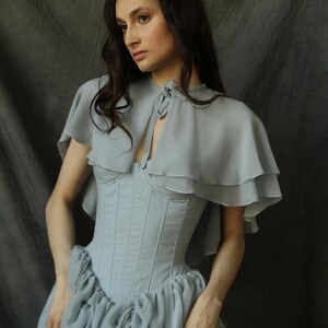 May include: A light blue, ruffled, sheer cape with a tied neckline, worn over a light blue corset and a gathered, light blue skirt.