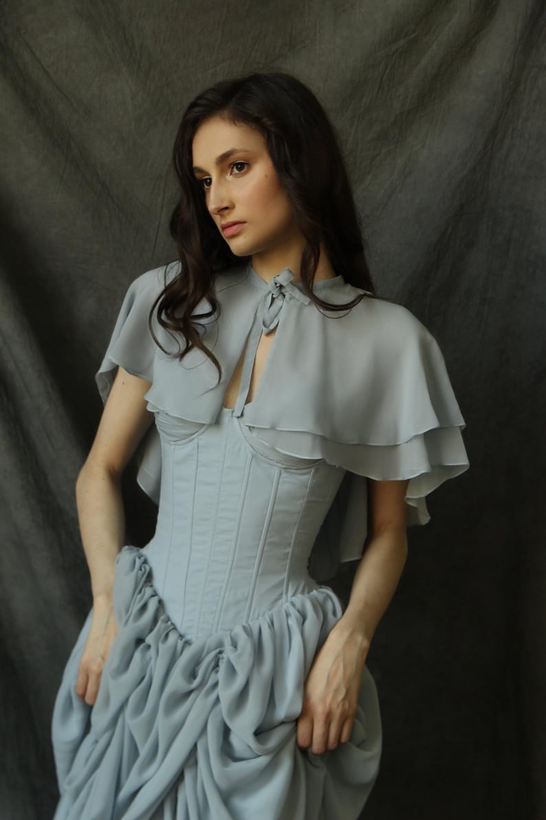 May include: A light blue, ruffled, sheer cape with a tied neckline, worn over a light blue corset and a gathered, light blue skirt.