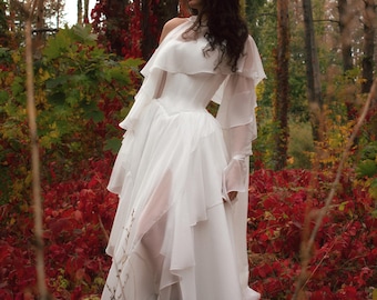 May include: A white, layered dress with a fitted bodice and flowing skirt. The dress features sheer, ruffled sleeves and a high neckline. The model is standing in a forest with red and green foliage.