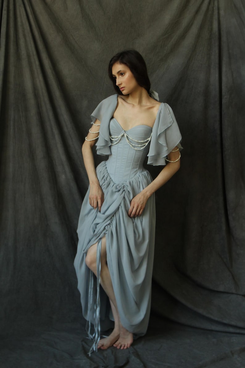 May include: A woman wearing a light blue dress with a ruffled bodice and pearl embellishments. The dress has a fitted bodice and a flowing skirt.