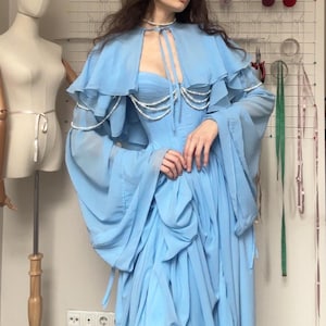 Op de afbeelding: Een lichtblauwe jurk met bijpassende cape en lange mouwen. De jurk heeft een gedrapeerd lijfje en rok, met parelaccenten. Een mannequin en naaimaterialen staan op de achtergrond.