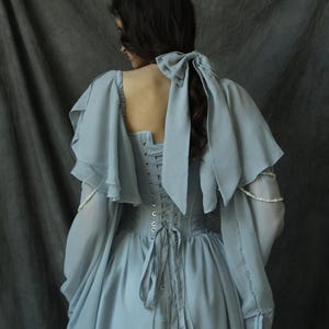 May include: A light blue dress with a lace-up back and a large bow at the back of the neck. The dress has long sleeves with ruffles at the shoulders and wrists.