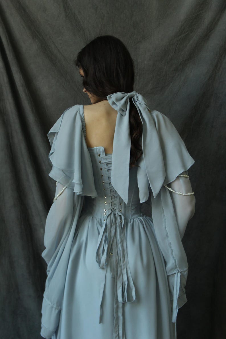 May include: A light blue dress with a lace-up back and a large bow at the back of the neck. The dress has long sleeves with ruffles at the shoulders and wrists.