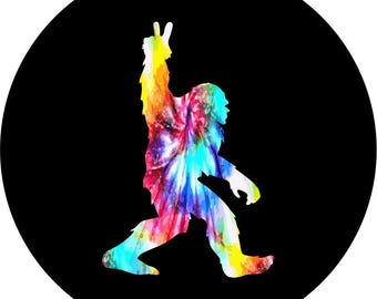 May include: A silhouette of a Bigfoot figure with a tie-dye pattern in shades of pink, blue, green, and yellow. The figure is standing with one leg raised and its arms outstretched.
