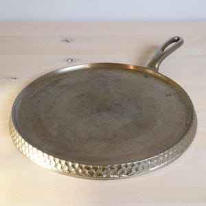 Op de afbeelding: Een ronde metalen bakplaat in antieke stijl met een handvat. De pan heeft een gehamerde textuur rond de rand en een glad kookoppervlak. De pan is gemaakt van een donker, metalen materiaal.