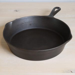 Pode incluir: Uma frigideira de ferro fundido preta com uma alça. A frigideira é redonda com uma lateral ligeiramente inclinada. A alça tem um orifício para pendurar. A frigideira está sobre uma superfície de madeira clara.