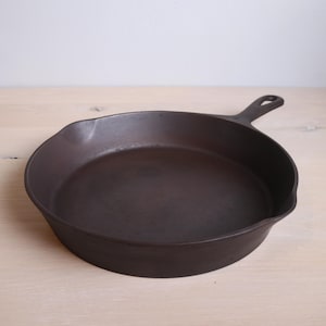 May include: A cast iron skillet with a black finish and a handle. The skillet is empty and sitting on a light wood surface.