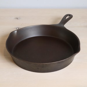 Op de afbeelding: Een donkerbruine gietijzeren koekenpan met een handvat en een afgerond kookoppervlak. De pan is gemaakt van zwaar metaal en is ontworpen om op een kookplaat of in een oven te koken. De pan is een klassiek keukengereedschap.