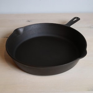 Op de afbeelding: Een zwarte gietijzeren koekenpan met een handvat en een schenktuit. De pan is rond en heeft een plat kookoppervlak. De handgreep heeft een gat om op te hangen. De pan staat op een lichtgekleurd houten oppervlak.