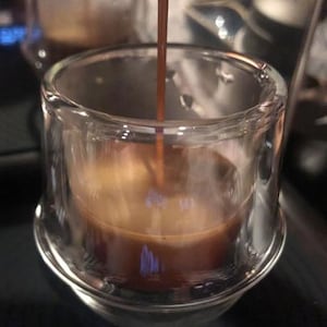 Puede incluir: Primer plano de una taza de vidrio transparente que se llena con caf&eacute; marr&oacute;n oscuro. El caf&eacute; se vierte desde un pico por encima de la taza.