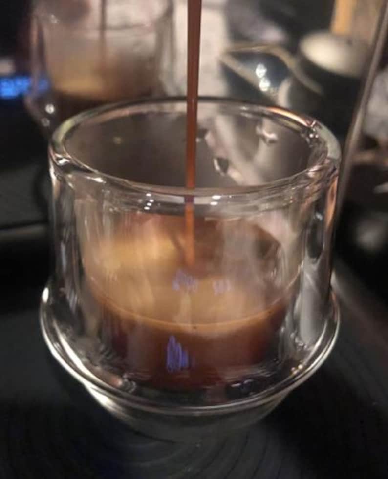 Puede incluir: Primer plano de una taza de vidrio transparente que se llena con caf&eacute; marr&oacute;n oscuro. El caf&eacute; se vierte desde un pico por encima de la taza.