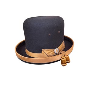 Op de afbeelding: Een zwarte wollen fedora hoed met een bruine leren band en een gouden lint. De hoed heeft een kleine gouden knop aan de voorkant en twee gouden kwastjes.