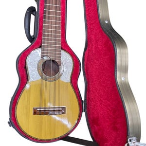 Könnte beinhalten: Ein hellbraunes Charango, ein kleines Anden-Saiteninstrument, liegt in seinem geöffneten Koffer. Das Charango hat eine perlmuttweiße Decke und ein dunkelbraunes Griffbrett. Der Koffer ist mit rotem Samt ausgekleidet.