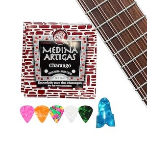Könnte beinhalten: Eine Packung Medina Artigas Charango-Saiten mit dem Text "Encordado para dos charangos" und "Set for two charangos". Außerdem eine Vielzahl bunter Gitarrenplektren und ein Teil eines Gitarrenhalses mit Saiten.