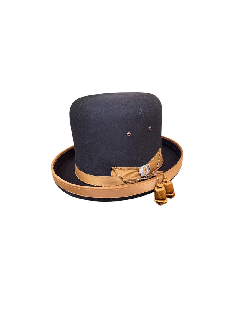 May include: A black felt fedora hat with a brown ribbon and two gold tassels. The hat has a small silver button on the front.