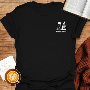 以下が含まれることがあります： 「Be Salt & Stay Lit」と「Matthew 5:13-14」のテキストが書かれた、2つの塩入れとろうそくの白いグラフィックが描かれた黒いTシャツ。