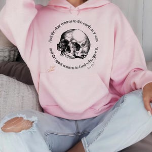 May include: A light pink hoodie with a black and white skull graphic. The graphic is surrounded by text that reads "And the dust returns to the earth as it was, and the spirit returns to God who gave it. Ecc 12:7."