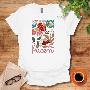 以下が含まれることがあります： 白いTシャツに花柄のデザインとピンク色の「Dona Nobis Pacem」の文字が書かれています。