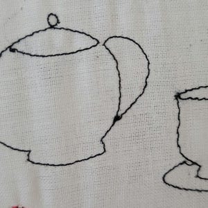 May include: Black and white embroidered image of a teapot and a teacup on a white fabric background.