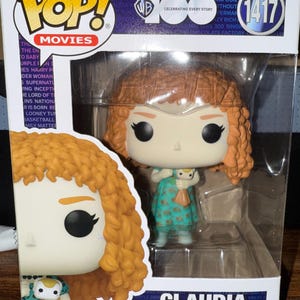 Könnte beinhalten: Funko Pop! Movies Figur von Claudia, einer Figur mit orangefarbenen Locken, die ein türkisfarbenes Kleid trägt und eine Puppe hält. Die Box zeigt das Funko Pop!-Logo, das Warner Bros.-Logo und die Nummer 1417.