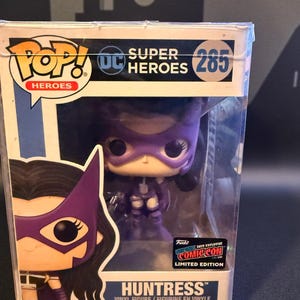 May include: A Funko Pop! vinyl figure of Huntress from DC Comics' Super Heroes series, in a clear display case. The box features the text "Huntress" and "285". It is a 2019 Comic-Con exclusive limited edition.