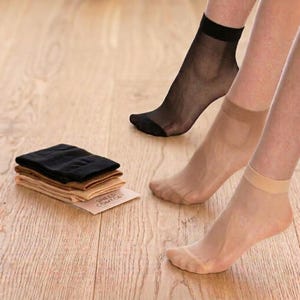 May include: A set of sheer ankle socks in black and various nude tones. One black sock is worn on a foot, and two nude-toned socks are worn on feet. A folded stack of matching socks rests on a card labeled Sheer Comfort.