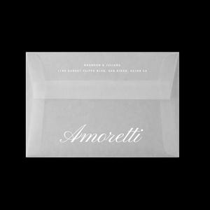 Puede incluir: Un sobre translúcido con la palabra "Amoretti" en escritura elegante. El sobre también presenta los nombres "Brandon & Juliana" y una dirección en texto blanco, sobre un fondo negro. El diseño es simple y sofisticado.