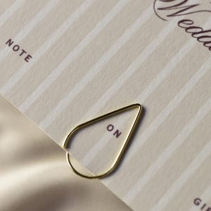 May include: A gold-colored paper clip in a teardrop shape, holding a cream-colored card with vertical stripes. The card has the word "NOTE" printed at the top left and the word "ON" printed on the paper clip.
