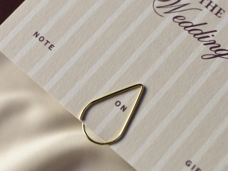 May include: A gold-colored paper clip in a teardrop shape, holding a cream-colored card with vertical stripes. The card has the word "NOTE" printed at the top left and the word "ON" printed on the paper clip.