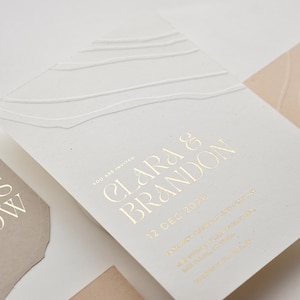 May include: White wedding invitation with gold foil lettering. The invitation reads "You are invited Clara & Brandon 12 Dec 2022".