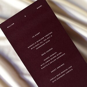 May include: A burgundy menu card with white text, featuring a list of dishes. The menu includes starters, main courses, and sweet endings. The names William and Naomi are at the top. The card is on a satin surface.