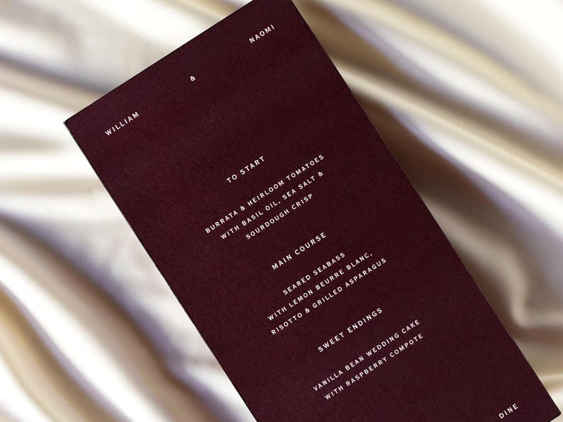 May include: A burgundy menu card with white text, featuring a list of dishes. The menu includes starters, main courses, and sweet endings. The names William and Naomi are at the top. The card is on a satin surface.