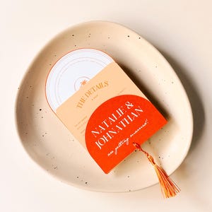 May include: A folded wedding invitation card in red and orange. The front of the card features a half-circle design with the text "Natalie & Jonathan are getting married!" The back of the card has the text "The Details" and a palm tree icon.